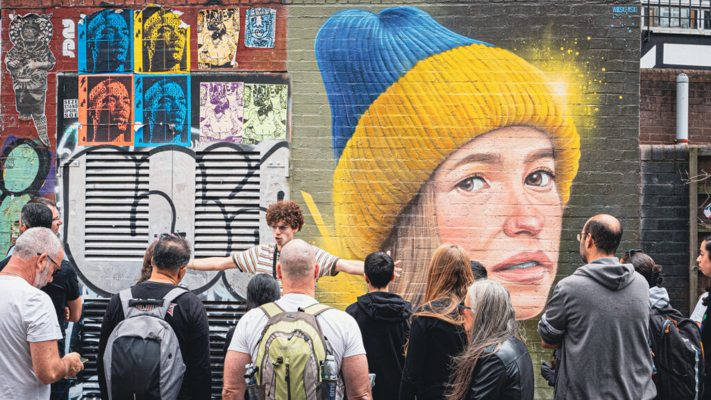 מדריך וקהל מטיילים עומדים על רקע ציור גרפיטי, סיור גרפיטי בשורדיץ'